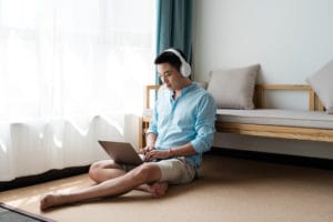 Young Asian man using laptop at home while wearing headphones. He is sitting on the floor on a tan rug.