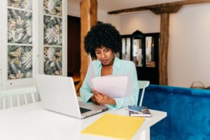 Young Black woman freelancer in casual outfit drinking coffee while sitting at table and working on project with laptop at home