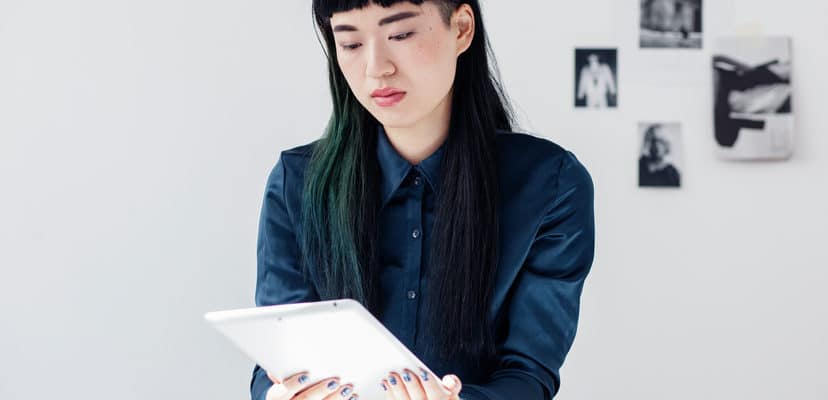 Portrait of a young asian woman working at a desk in a modern studio office. She has a focused confident expression and is looking down towards a tablet she is holding. She has long black hair with a green streak and bangs. She is wearing a blue shirt and leaning on the desk.