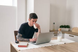 Short haired man working with laptop in kitchen while drinking milk