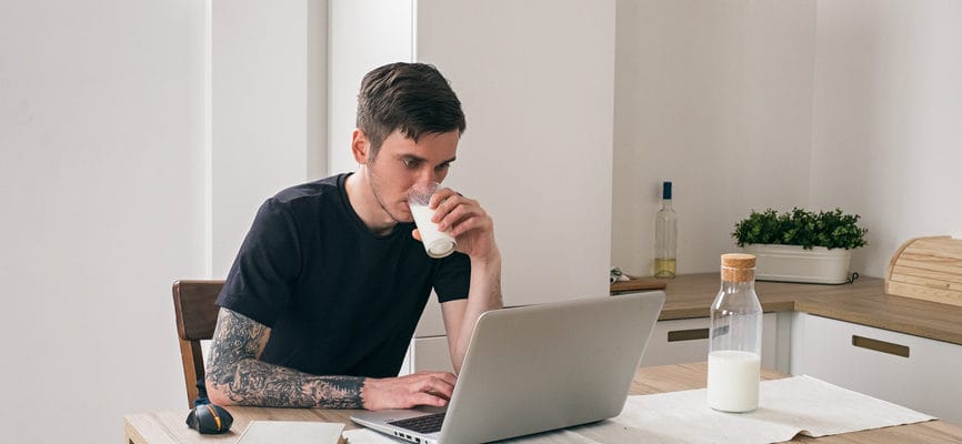 Short haired man working with laptop in kitchen while drinking milk