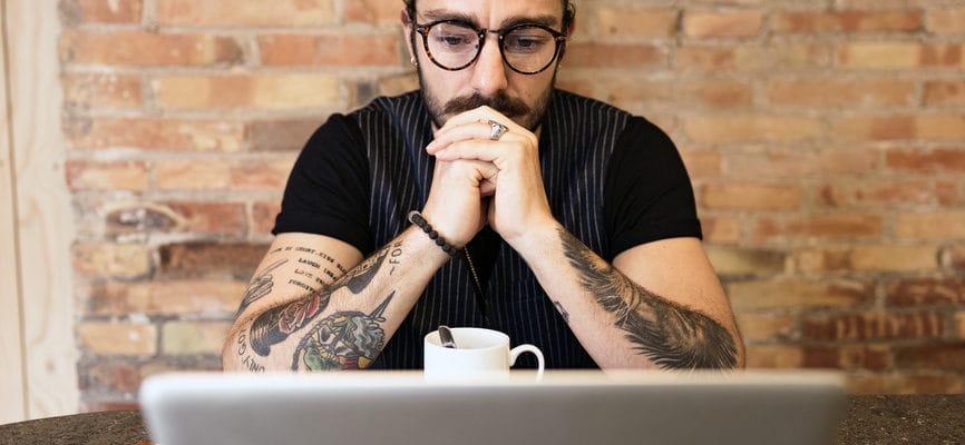 Portrait of good-looking stylish man with beard and tattoos in round glasses looking pensively at laptop with hands at face. He is sitting in cafe against brick wall.