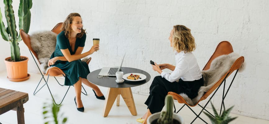 Two white women sitting together at a round black table laughing and drinking coffee.