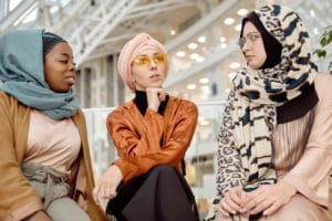 Three young muslim women sitting on next to each other. Women on the left is wearing a light blue head scarf with brown jacket, woman in the middle wearing a light pink headwrap with burnt orange top, and woman on the right wearing animal print headscarf with a light pink top.