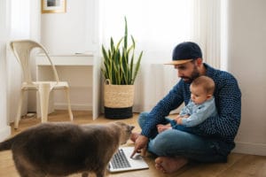 Father wearing baseball cap holding baby in arms while sitting on floor with his laptop on the ground. Their pet cat is walking by on the left bottom right corner of photo.