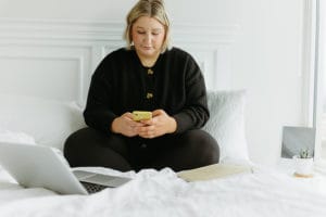Woman sitting cross legged on bed while using phone with yellow phone case and laptop is open next to her
