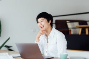 Asian woman wearing a white shirt smiling while working on her laptop. Mint green coffee mug on table