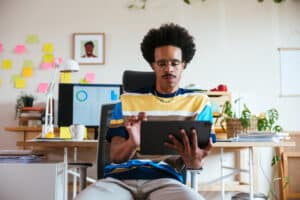 Focused mixed race man in casual clothes and glasses browsing and reading data on tablet while sitting on chair and working remotely in home office.