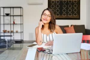 Smiling young woman using a laptop at a table in her living room while working remotely from home
