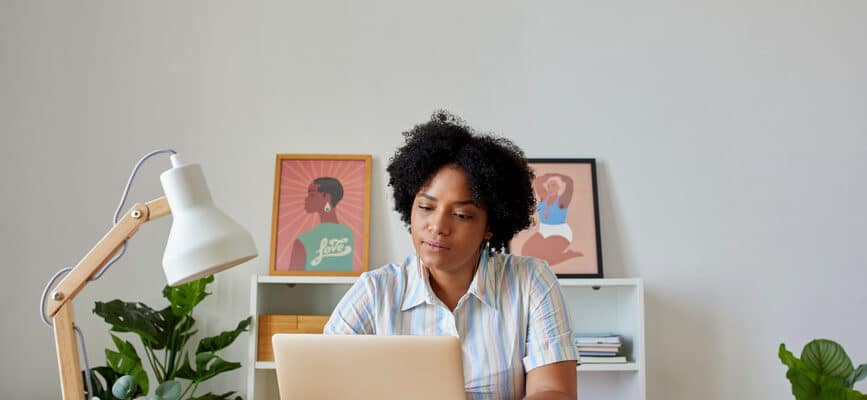 Curly haired African American female freelancer browsing netbook while working on remote project at table in modern home office