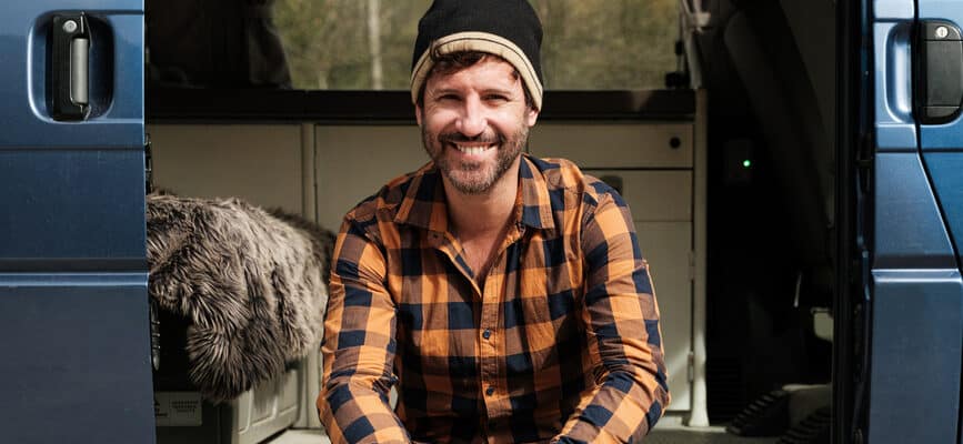 Portrait of cheerful man with winter hat looking at camera and smiling, sitting in his caravan during a road trip in autumn holding a laptop